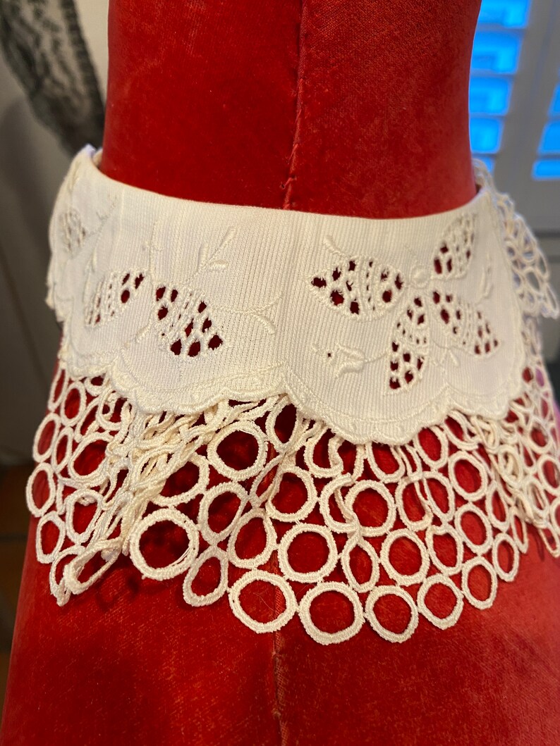 Puede incluir: Collar de encaje blanco con borde festoneado y delicado bordado floral. El collar est&aacute; sobre un fondo de terciopelo rojo.
