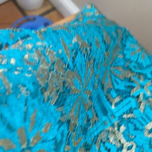 May include: Turquoise blue lace fabric with a gold metallic pattern. The lace has a delicate floral design.