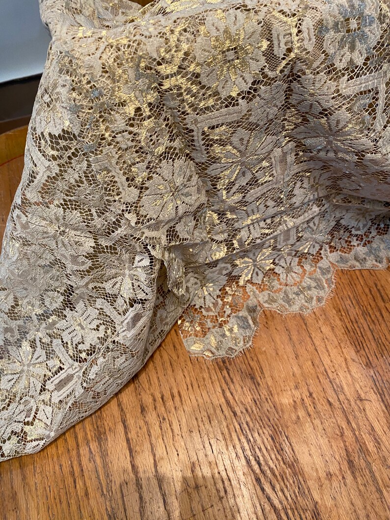 May include: Beige lace fabric with a gold metallic pattern. The lace has a delicate floral design.