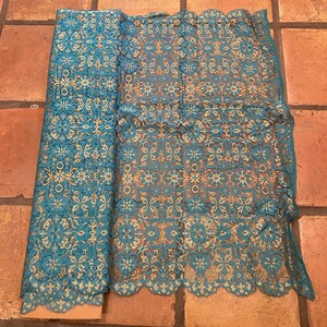 May include: Teal and gold lace fabric with a geometric pattern. The fabric is rolled up and laid flat on a terracotta tile floor.