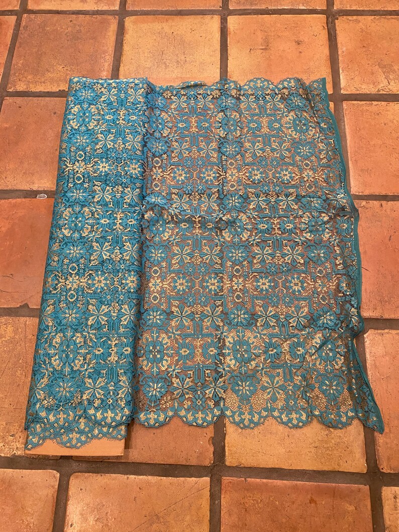 May include: Teal and gold lace fabric with a geometric pattern. The fabric is rolled up and laid flat on a terracotta tile floor.