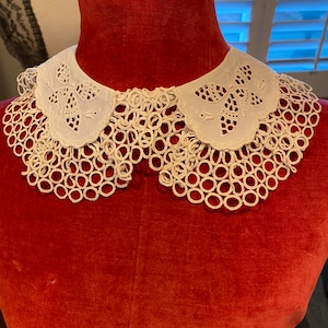 Puede incluir: Un cuello de encaje blanco con borde festoneado y bordado delicado. El cuello se muestra en un maniqu&iacute; de terciopelo rojo.