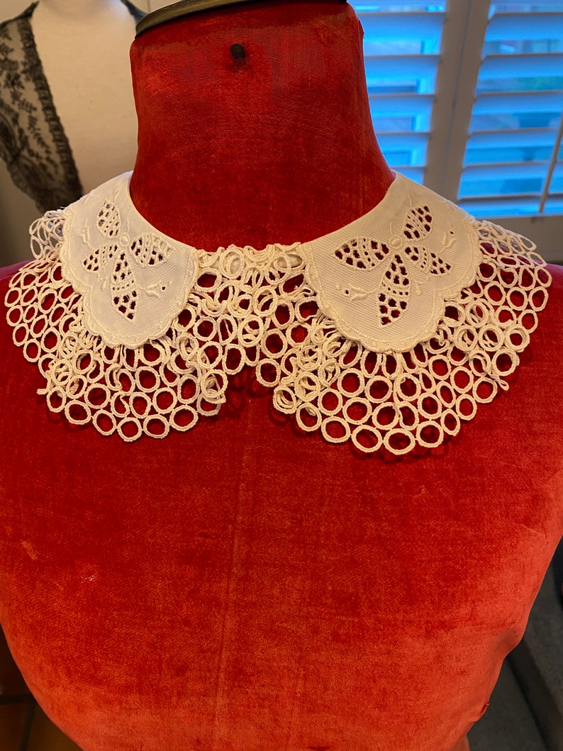 Puede incluir: Un cuello de encaje blanco con borde festoneado y bordado delicado. El cuello se muestra en un maniqu&iacute; de terciopelo rojo.