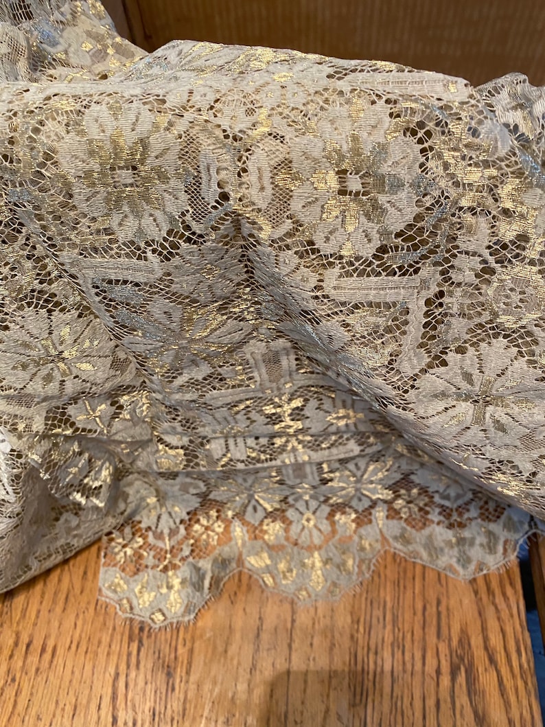 May include: A close-up of a beige lace fabric with gold metallic accents. The lace has a delicate floral pattern and a scalloped edge.