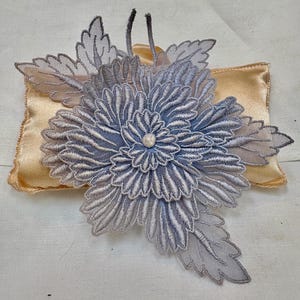May include: A layered, light blue embroidered floral applique featuring intricate petal stitching and a central pearl-like bead. The flower is positioned atop a rectangular, smooth, gold-colored satin cushion with stitched edges and a soft sheen.