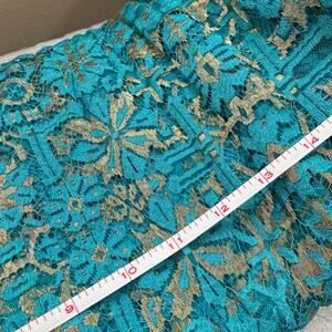 May include: Teal lace fabric with gold embroidery. A white measuring tape is laid across the fabric, showing measurements from 8 to 14 inches.
