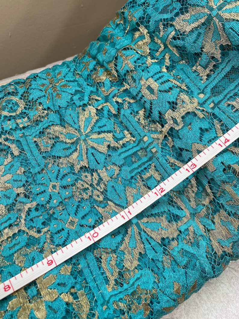 May include: Teal lace fabric with gold embroidery. A white measuring tape is laid across the fabric, showing measurements from 8 to 14 inches.