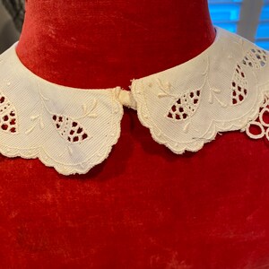 Puede incluir: Un cuello de encaje blanco con borde festoneado. El cuello est&aacute; sobre un maniqu&iacute; de terciopelo rojo.