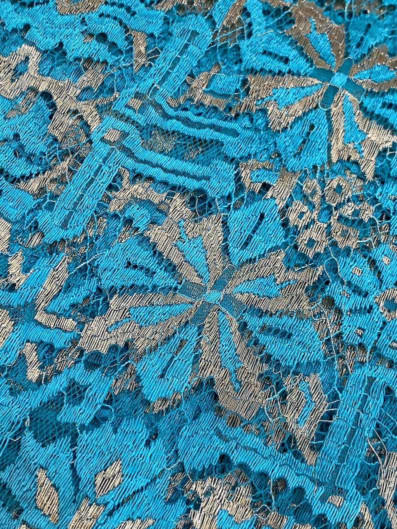 May include: Close-up of a turquoise lace fabric with a silver metallic pattern. The lace has a delicate floral design.