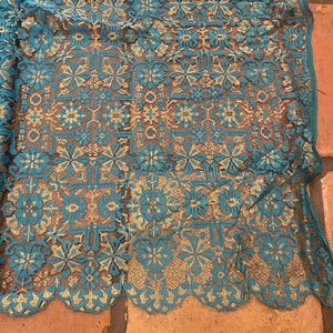 May include: Teal lace fabric with a gold pattern of flowers and geometric shapes. The lace has a scalloped edge.