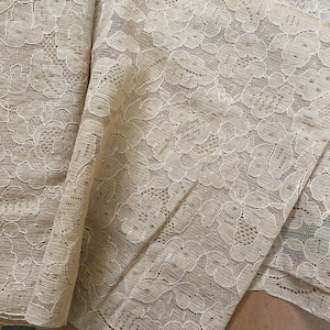 Könnte beinhalten: Ein beigefarbener Spitzenstoff mit einem Blumenmuster. Der Stoff ist gefaltet und auf einer braunen Oberfläche ausgelegt.