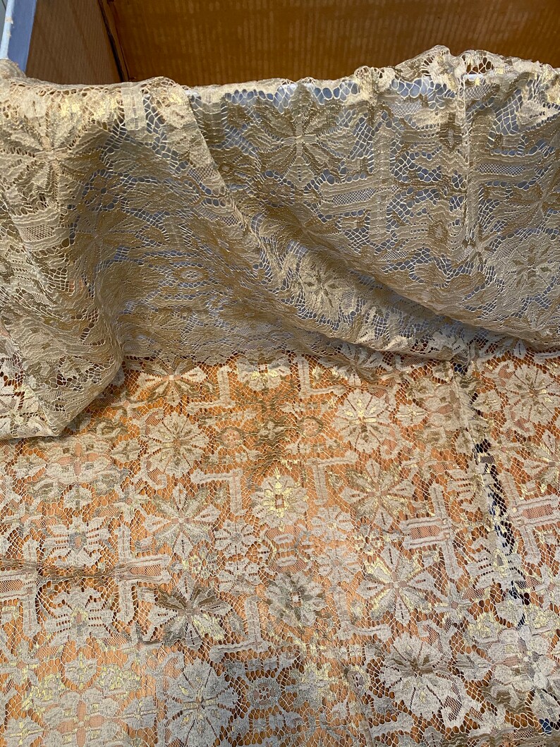 May include: A close-up of a gold lace fabric with a delicate floral pattern. The fabric is soft and sheer, and it has a subtle shimmer.