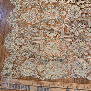 May include: A close-up of beige lace fabric with a gold metallic pattern. The fabric has a scalloped edge and a repeating floral design. A wooden ruler with the text "Lachman Bros. One of America's Largest Home Furnishers" is visible at the bottom of the image.