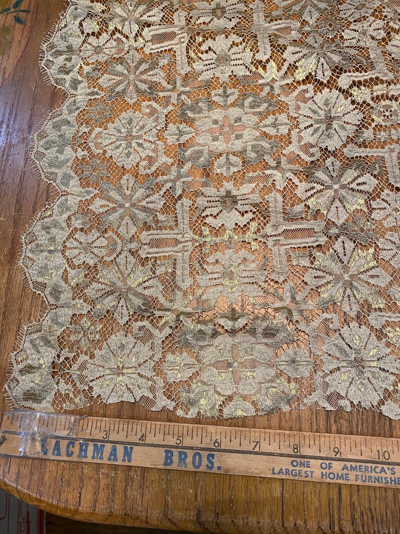 May include: A close-up of beige lace fabric with a gold metallic pattern. The fabric has a scalloped edge and a repeating floral design. A wooden ruler with the text "Lachman Bros. One of America's Largest Home Furnishers" is visible at the bottom of the image.