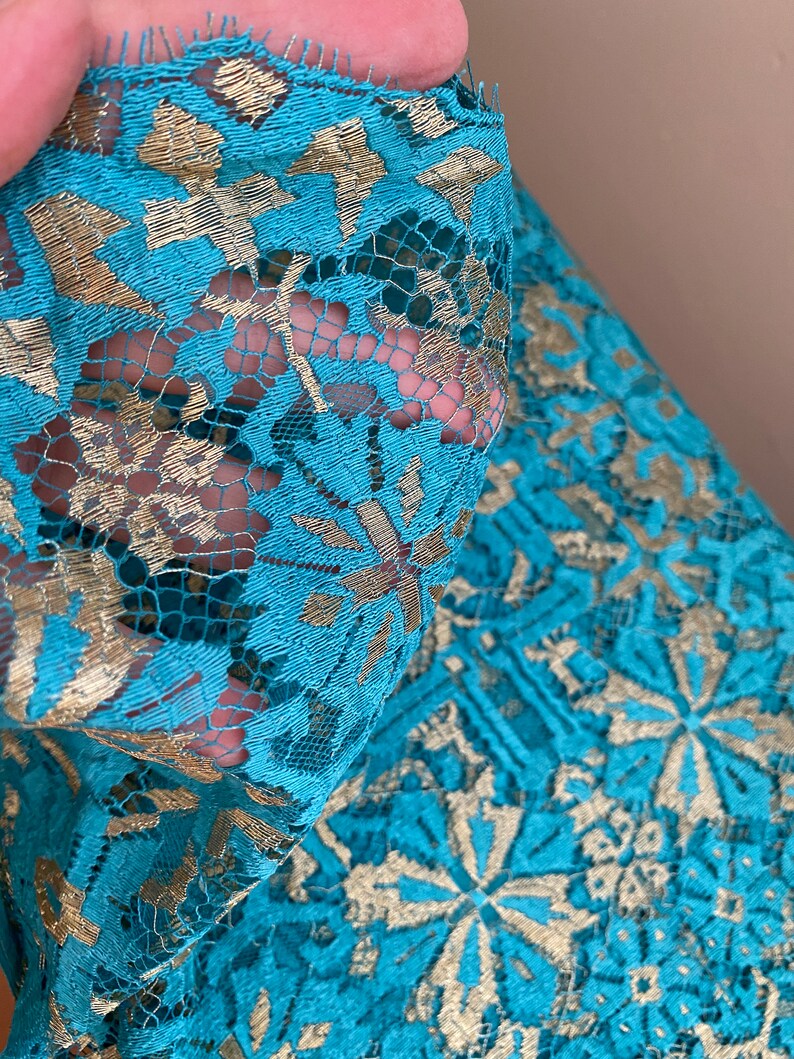 May include: Teal lace fabric with a gold metallic pattern. The lace has a delicate floral design.