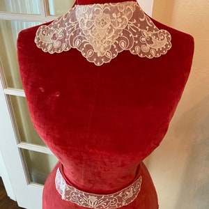 May include: A red velvet dress form with two white lace collars. The top collar is a wide, scalloped collar with a floral design. The bottom collar is a narrower band with a similar floral design.