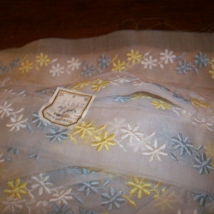 Könnte beinhalten: Weißer, durchsichtiger Stoff mit gestickten Blumenmustern in Gelb, Blau und Weiß. Der Stoff hat ein Etikett mit der Aufschrift "Clayson Fabrics Made in Switzerland".