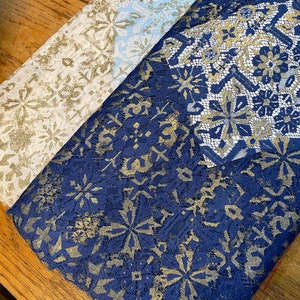 May include: Two pieces of fabric with a gold and blue lace pattern. The fabric on the left is a light beige color and the fabric on the right is a dark blue color.