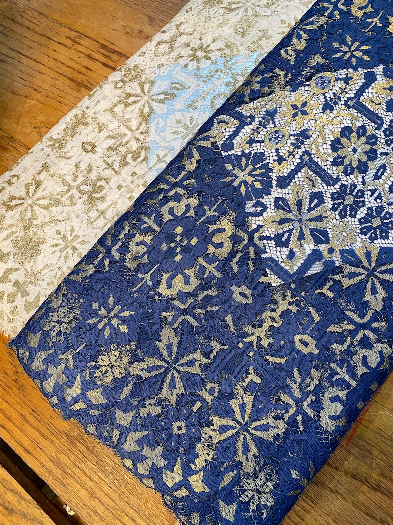 May include: Two pieces of fabric with a gold and blue lace pattern. The fabric on the left is a light beige color and the fabric on the right is a dark blue color.