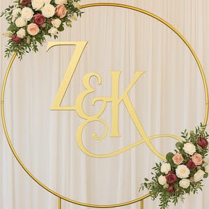 May include: A gold metal wedding arch with the initials Z & K. The arch is decorated with floral arrangements of white, pink, and burgundy roses. The background is a white curtain.