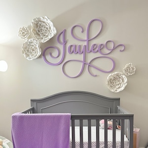 May include: A purple and white nursery with a crib, a baby swing, and a rug. The crib has a purple blanket with the name "Jaylee Lynne" embroidered on it. The wall is decorated with a purple wooden sign that says "Jaylee" and white paper flowers.