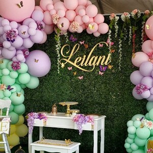 May include: A first birthday party decoration with a green backdrop, pastel balloons, a white table with a cake stand, and a white high chair with a banner that says "ONE". The backdrop is decorated with flowers and butterflies. The name "Leilani" is written in gold letters on the backdrop.