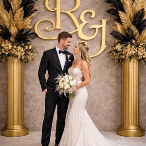 May include: A wedding scene featuring a couple in formal attire. The bride wears a white, sequined mermaid gown and holds a bouquet. The groom is in a black tuxedo. Gold pillars and floral arrangements frame the couple, with a gold monogram backdrop.