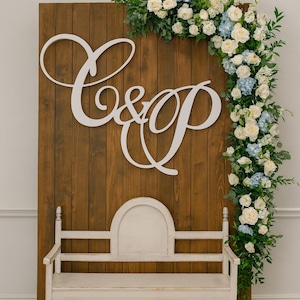 May include: A wooden backdrop with the letters "C&P" in white script, adorned with a floral arrangement of white roses, blue hydrangeas, and greenery. A white bench sits in front of the backdrop, creating a charming wedding or event setting.