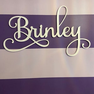 Może przedstawiać: Biały drewniany znak z napisem "Brinkley" napisanym kursywą. Znak znajduje się na ścianie w pasy fioletowe i jasnofioletowe.