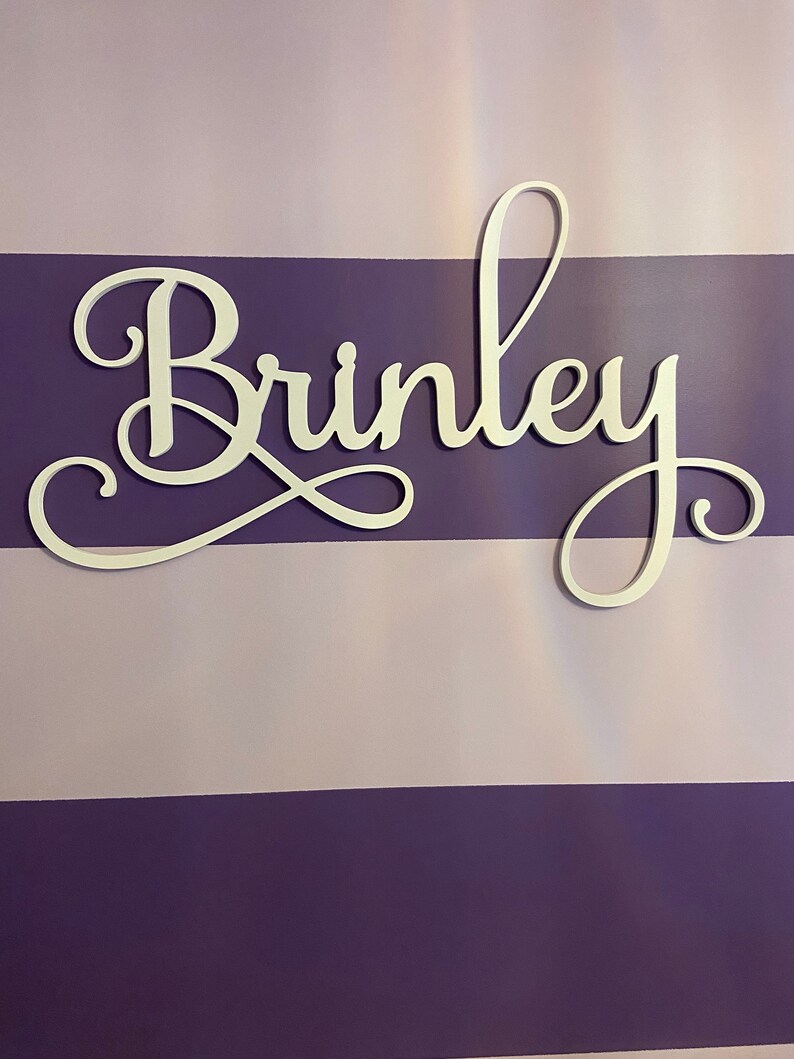 Może przedstawiać: Biały drewniany znak z napisem "Brinkley" napisanym kursywą. Znak znajduje się na ścianie w pasy fioletowe i jasnofioletowe.