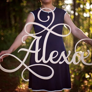 May include: A white wooden sign with the name "Alexia" written in a cursive font. The sign is held by a person in front of a blurred background of green trees.