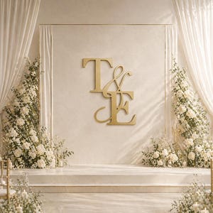 May include: A wedding ceremony backdrop featuring a gold monogram of the letters "T & E" against a cream-colored wall. White floral arrangements and draped curtains frame the stage. Gold chairs are visible in the foreground.