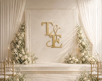 May include: A wedding ceremony backdrop featuring a gold monogram of the letters "T & E" against a cream-colored wall. White floral arrangements and draped curtains frame the stage. Gold chairs are visible in the foreground.
