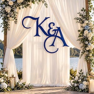 May include: A wooden wedding arch draped with white fabric, featuring a large blue monogram of the letters K&A. The structure is decorated with white and light blue floral arrangements and greenery, with gold lanterns containing candles at the base.