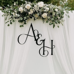 May include: A white backdrop with sheer fabric and a floral arrangement of white roses and greenery. Black cursive letters "A & H" are centered on the backdrop, suitable for a wedding or event.