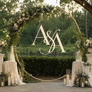 May include: A large circular gold metal wedding arch decorated with white flowers and greenery, featuring the letters A&A in the center. The arch is flanked by two white fluted pedestals topped with floral arrangements and white pillar candles.