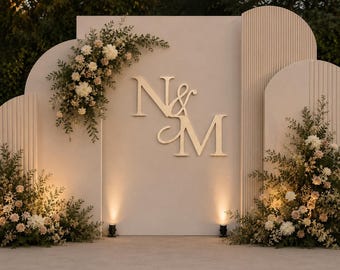 May include: A beige event backdrop featuring three arched panels with vertical ribbed textures. The center panel displays large, gold-colored letters N & M. Floral arrangements of white and pale pink flowers with green foliage are placed on the left and right.