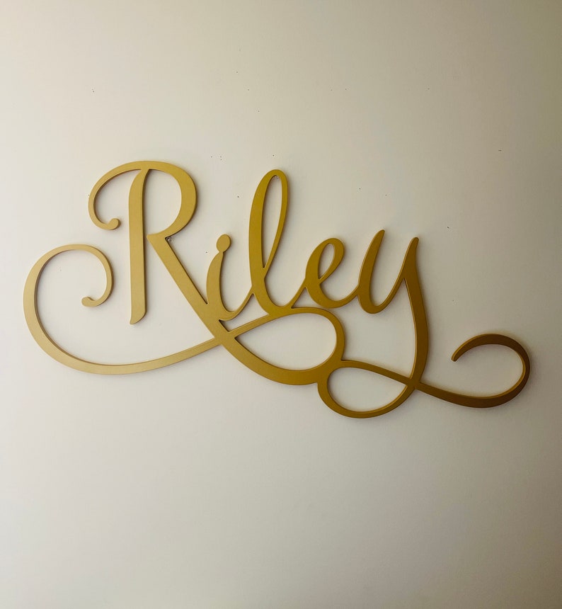 Może przedstawiać: Złoty znak z imieniem w ozdobnym, ozdobnym stylu pisma odręcznego z imieniem "Riley".