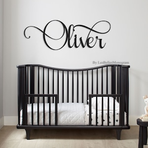 May include: Black metal cursive name sign with the name "Oliver" in a decorative font. The sign is mounted on a white wall above a black crib with a white mattress and a white and black star patterned sheet.