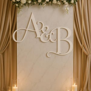 May include: A wedding backdrop featuring a gold frame, a marble-look panel with the letters "A & B", and a floral arrangement of white flowers. Lit candles in glass cylinders are at the base. Beige curtains frame the backdrop.