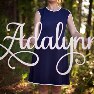 May include: A large, cursive wooden sign spells out the name "Adalynn". The sign is held in front of a person wearing a navy blue dress with white trim. The background is a blurred view of a green forest.