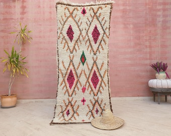 May include: A white Moroccan rug with a diamond pattern in shades of pink, orange, and green. The rug is hanging on a pink wall.