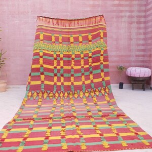 May include: A colorful Moroccan rug with a geometric pattern in shades of red, yellow, and green. The rug has a fringed edge and is woven with a textured, hand-knotted style.