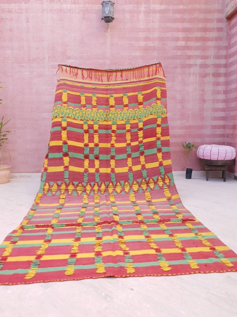 May include: A colorful Moroccan rug with a geometric pattern in shades of red, yellow, and green. The rug has a fringed edge and is woven with a textured, hand-knotted style.