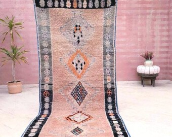 May include: A rectangular, peach-colored rug with geometric patterns in black, orange, and blue. The rug has a black border with white and black circular designs. The rug is displayed against a pink wall.