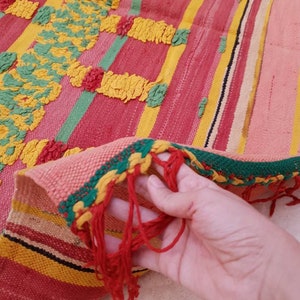 May include: A colorful woven rug with a fringe. The rug has stripes of red, yellow, green, and pink. The rug is made with a textured weave.
