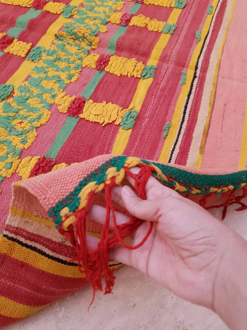 May include: A colorful woven rug with a fringe. The rug has stripes of red, yellow, green, and pink. The rug is made with a textured weave.