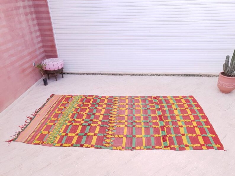 May include: A red, yellow, and green woven rug with a checkered pattern. The rug is laid out on a white tiled floor.