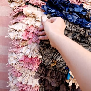 Puede incluir: Una alfombra colorida tejida a mano con retazos de tela reciclados. La alfombra presenta una variedad de colores, incluyendo rosa, azul, blanco y gris. La alfombra est&aacute; hecha de muchas piezas peque&ntilde;as de tela individuales que se han tejido juntas para crear una superficie &uacute;nica y texturizada.