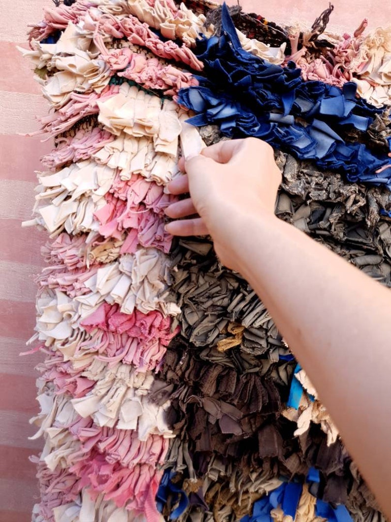 Puede incluir: Una alfombra colorida tejida a mano con retazos de tela reciclados. La alfombra presenta una variedad de colores, incluyendo rosa, azul, blanco y gris. La alfombra est&aacute; hecha de muchas piezas peque&ntilde;as de tela individuales que se han tejido juntas para crear una superficie &uacute;nica y texturizada.
